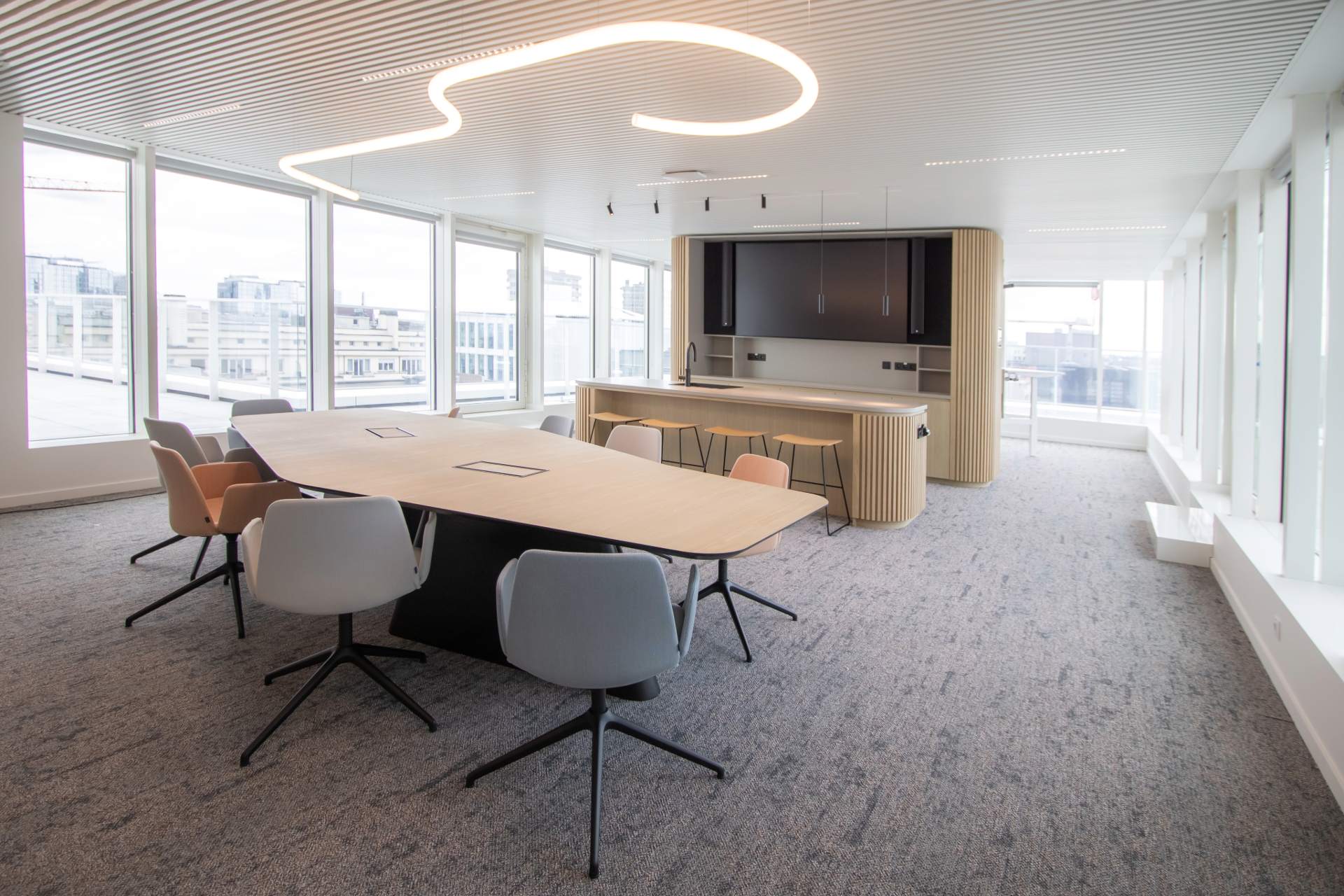 Grande pièce avec espace cuisine et grande tables en bois clair, moquette grise, finition bois et métal noir