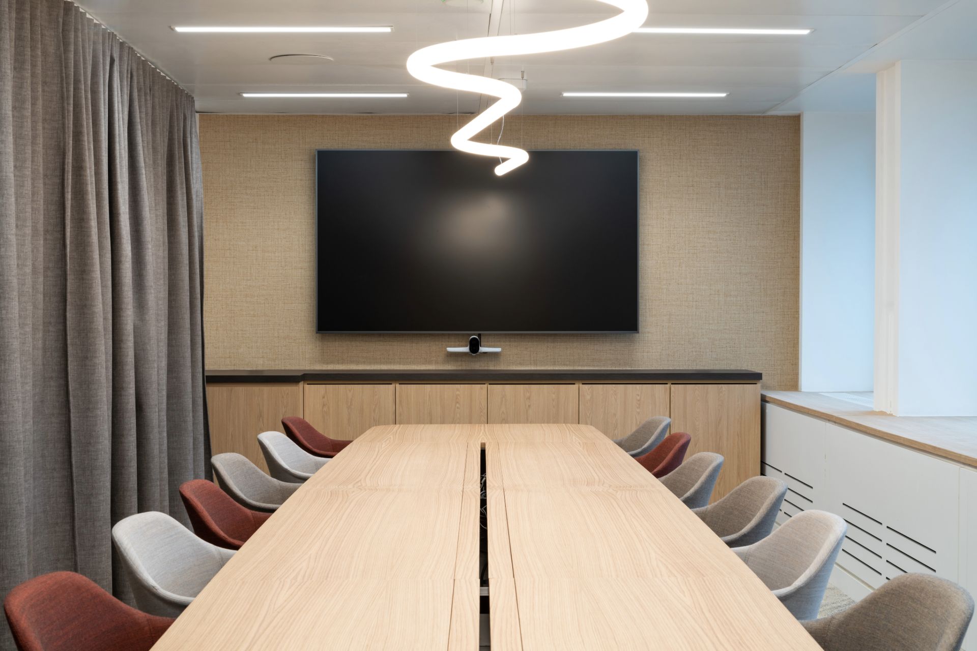 Grande salle de réunion avec un rideau pour la confidentialité, un luminaire formant une ligne qui ondule est au dessus de la grande table.