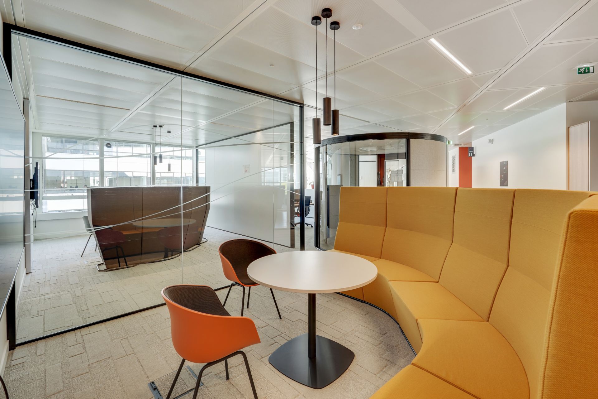 Espace de réunion informelle, banquette en demi lune couleur ôcre, chaises rouge et table en bois clair