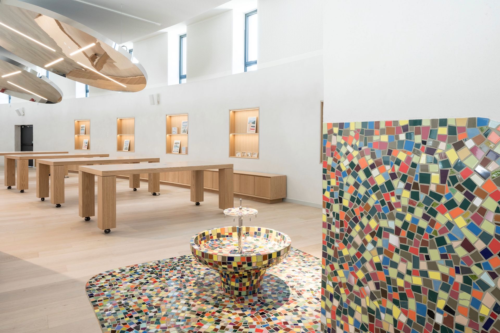 A l'étage, tables en bois clair devant les étagères avec des pièces exposées, et au premier plan une fontaine en mosaïque multicolore - Maison des métiers d'art