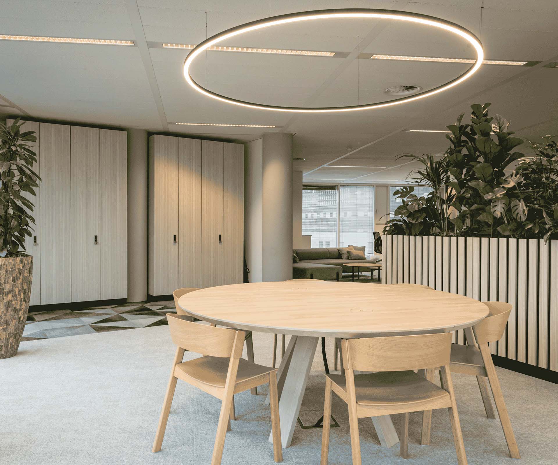 Espace de coworking ou pour des réunions informelles avec chaises et table en bois clair. Une luminaire en forme de cercle est au dessus de la table. L'ensemble est dans les tons clairs beige et gris.