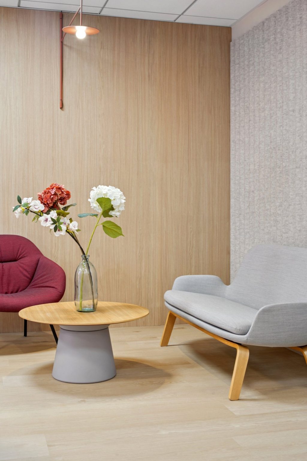 Photo avec un canapé gris et un fauteuil rouge avec au centre une table basse en bois surmontée par des fleurs dans une bouteille en verre.