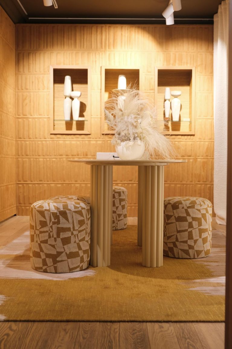 Décor en bois clair, table ronde au centre de la pièce avec bouquet de pampa, entourée de ses fauteuils en tissus marrons et beige. Décorations encastrées dans le mur