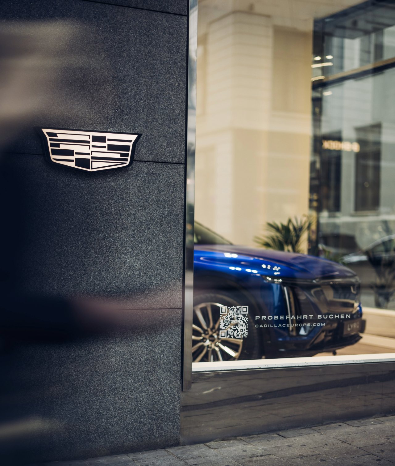 Façade extérieure avec le logo Cadillac à gauche et un véhicule bleu derrière une vitre à droite.