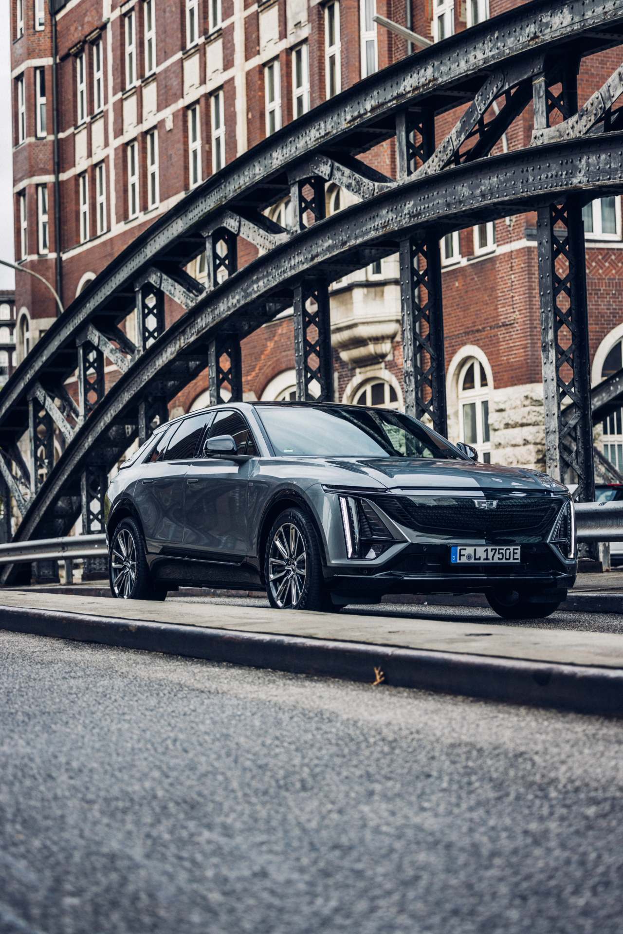 Cadillac grise dans une rue avec un pont métallique devant un immeuble en brique.