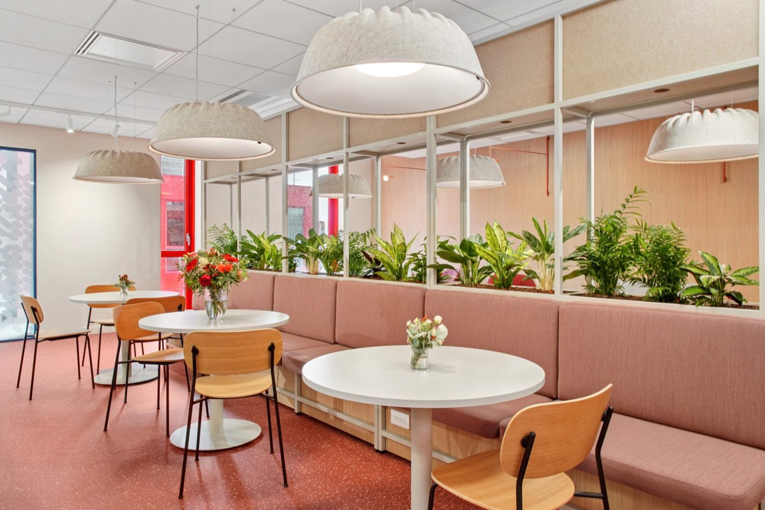 Photo of a dining room with three tables, chairs, and a bench. Plants form a partition between this area and the other side.