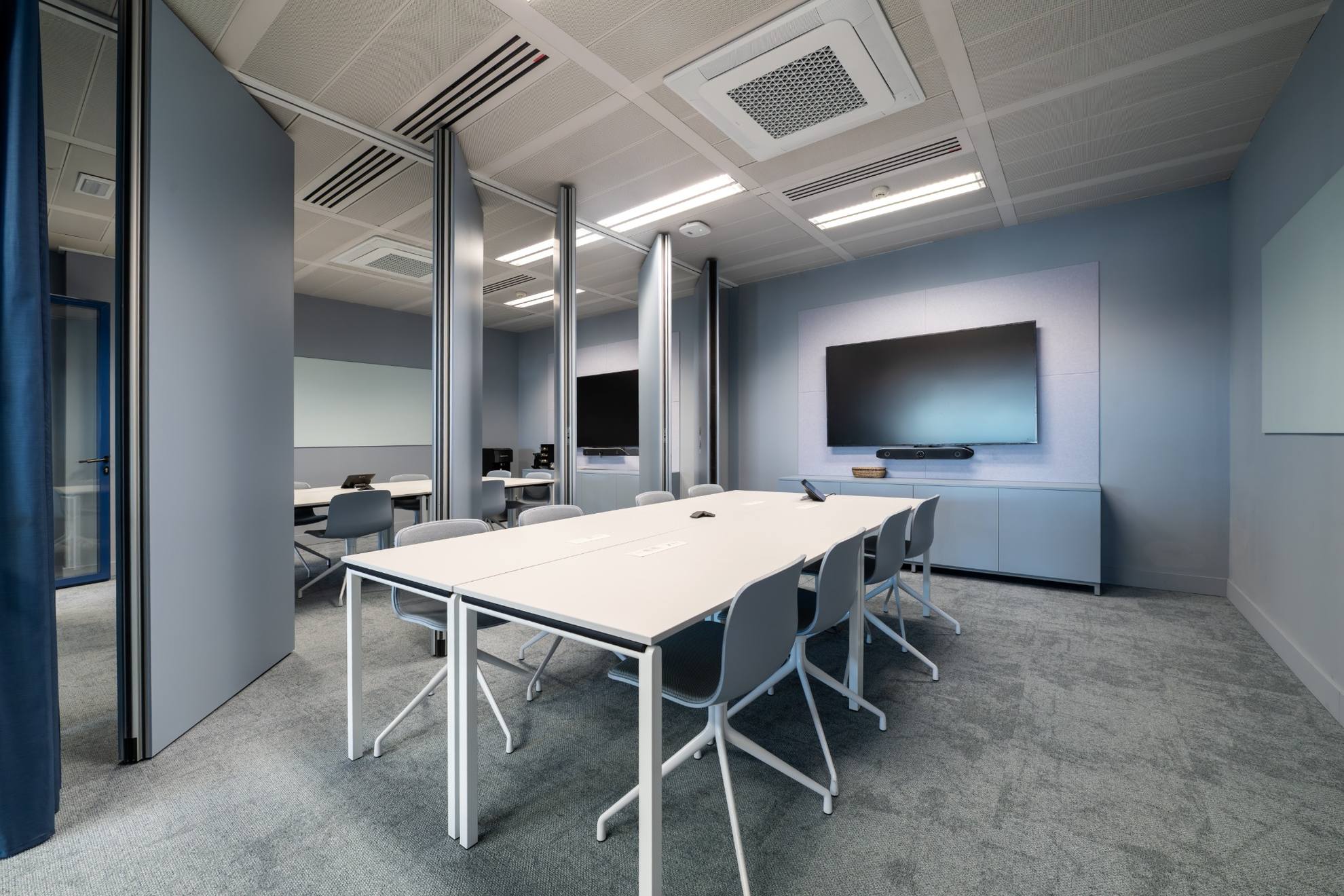 Salle de réunion gris avec une table, 8chaises et une télévision murale.