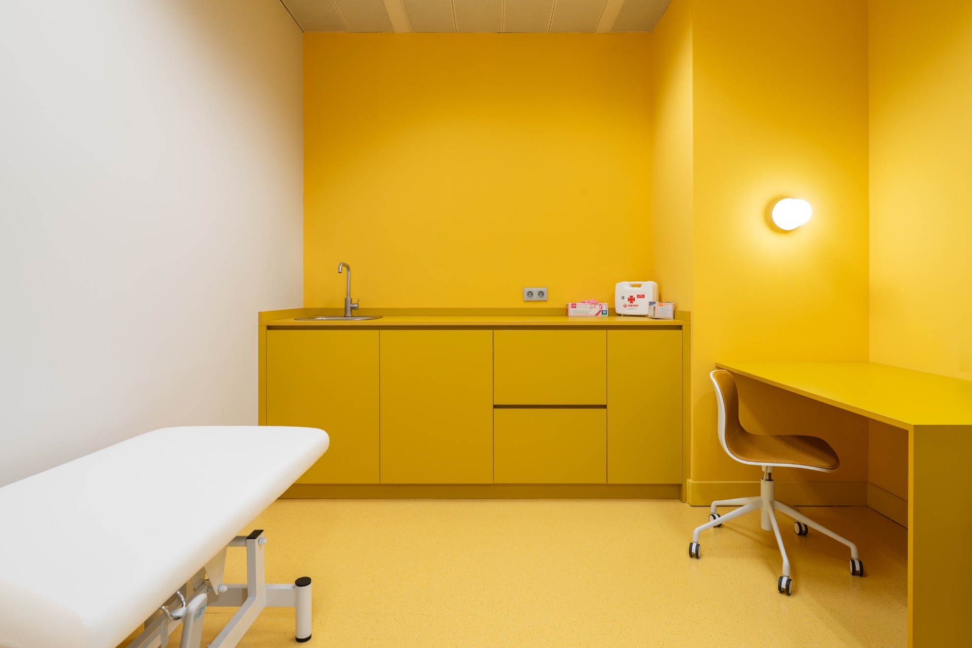 Salle médicale avec murs jaunes un bureau, un lit plus une planche de travail.