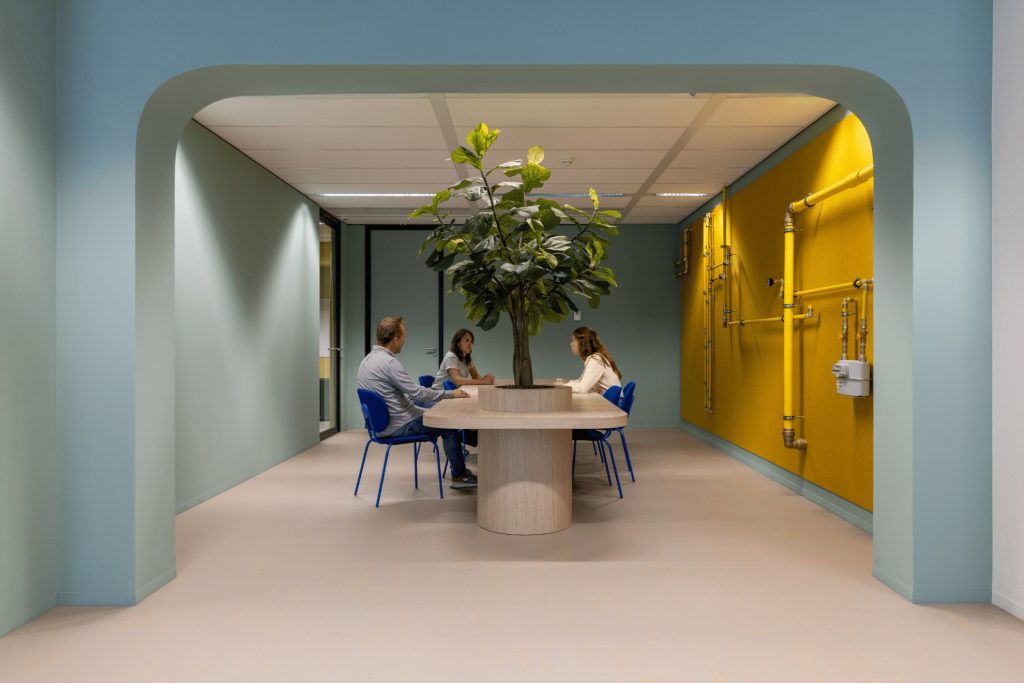 Une salle de réunion avec un arbre au centre et des murs bleus avec des tuyaus jaunes à droite.