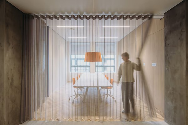 Une salle de réunion avec 6 chaises orange et une lampe suspendue orange avec une personne tirant des rideaux.