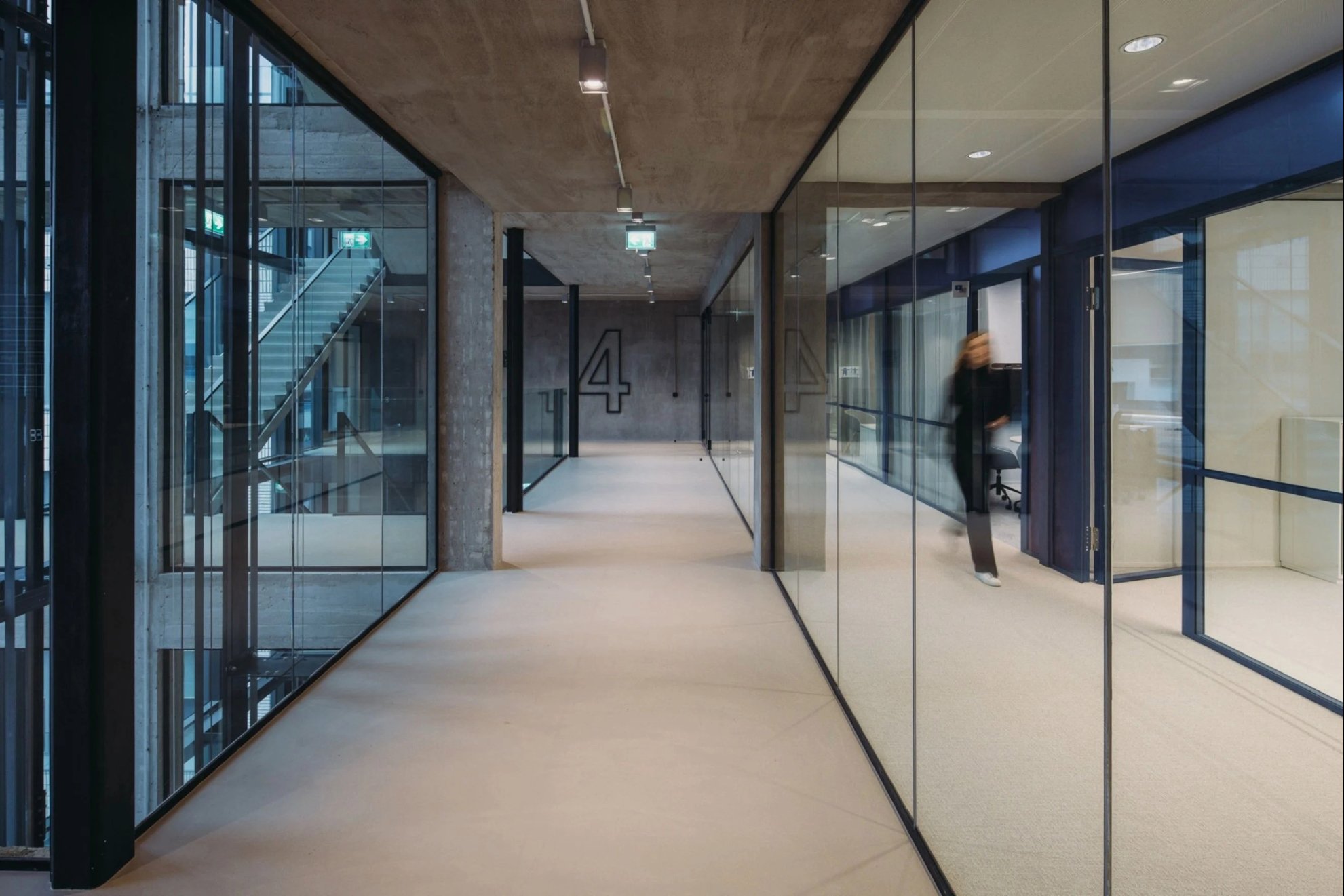 Un couloir avec le chiffre 4 sur le mur brut, à gauche des escaliers et à droite des vitres avec une femme marchant.