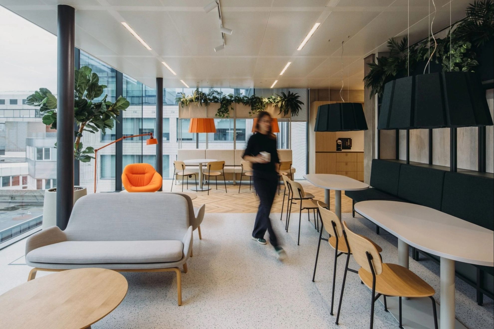 Image d'une cafétéria avec des tables et des chaises, une femme marchant avec un verre et à droite des vitres donnant sur des autres bâtiments.