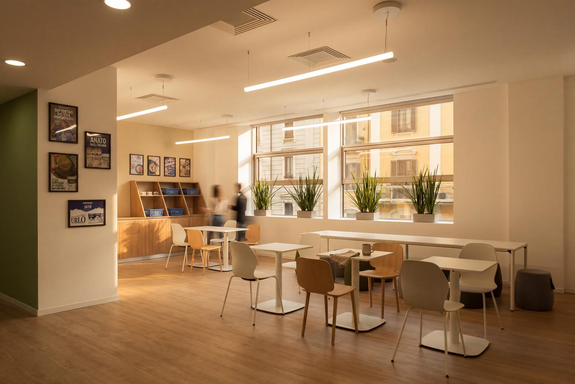 Une cafétéria ou zone de détente lumineuse avec des tables blanches, des chaises en bois et des plantes vertes alignées devant les fenêtres.