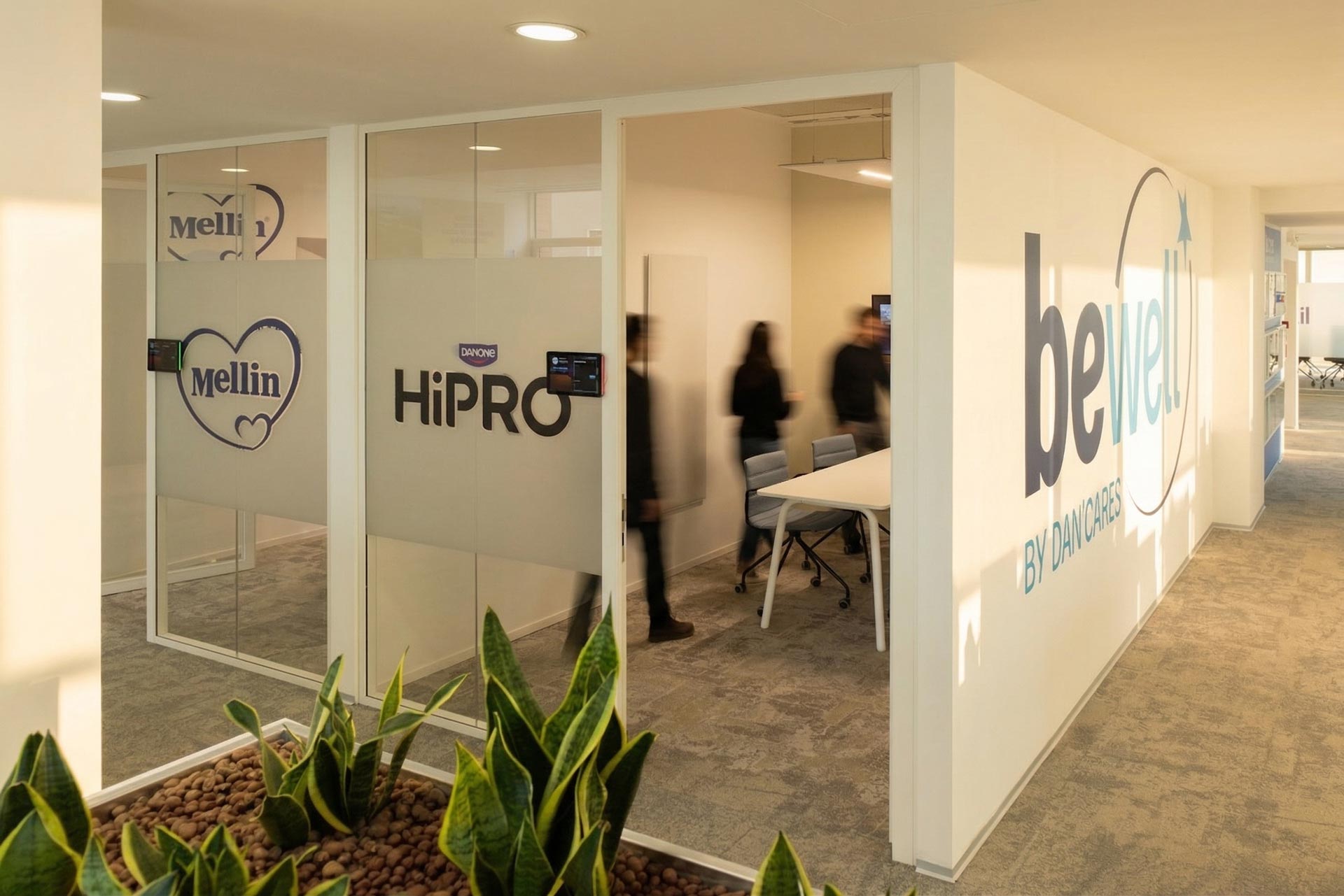Un couloir desservant plusieurs salles de réunion vitrées aux noms de marques (Mellin, HiPRO) avec un mur graphique "be well".