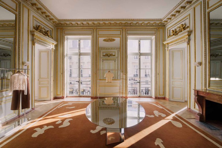 Un grand salon de réception prestigieux aux détails baroques et dorés, transformé en espace d'exposition avec une table miroir centrale et des portants à vêtements.