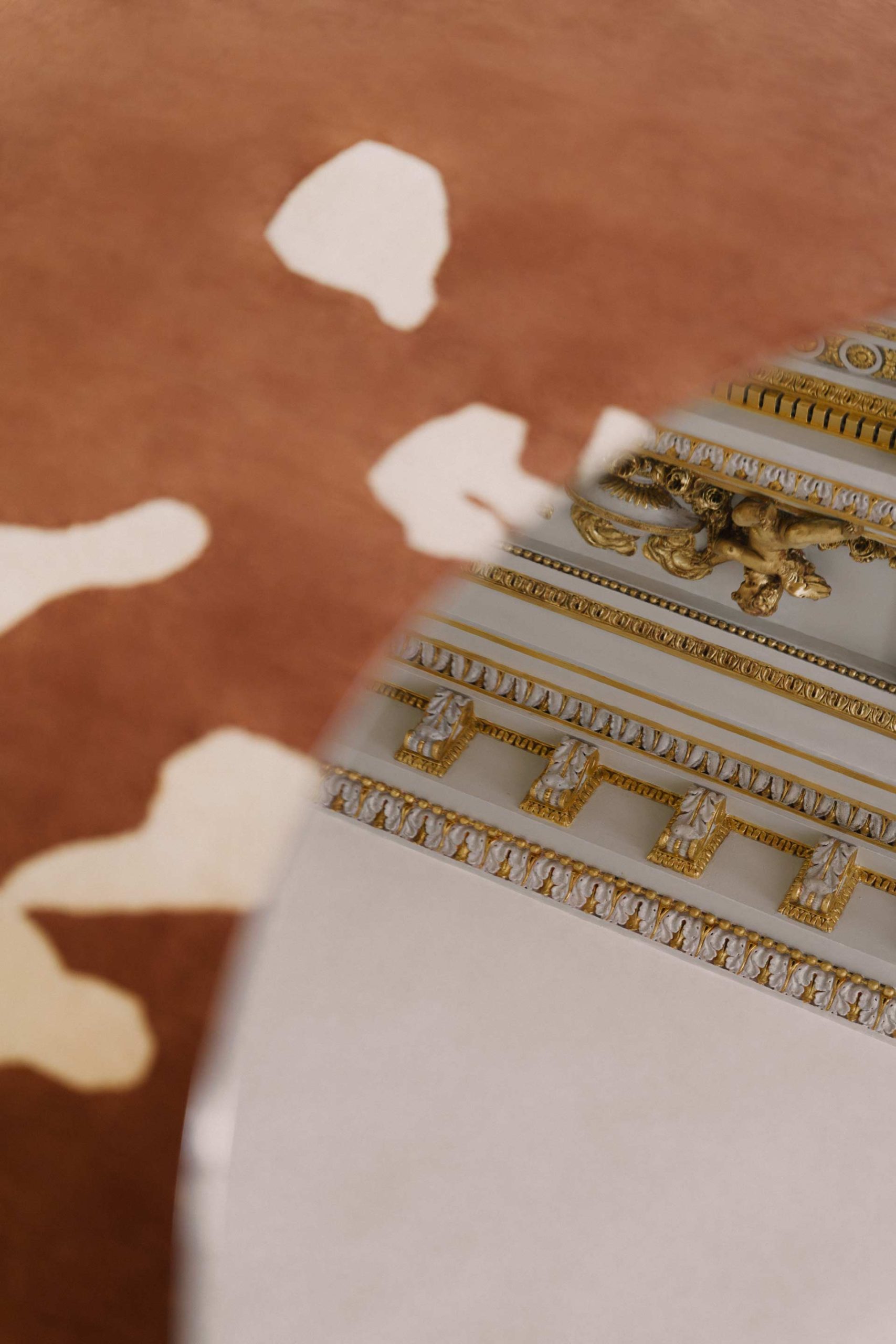 Un reflet artistique dans une surface miroir montrant les détails sculptés et dorés d'un plafond classique contrastant avec un tapis brun.