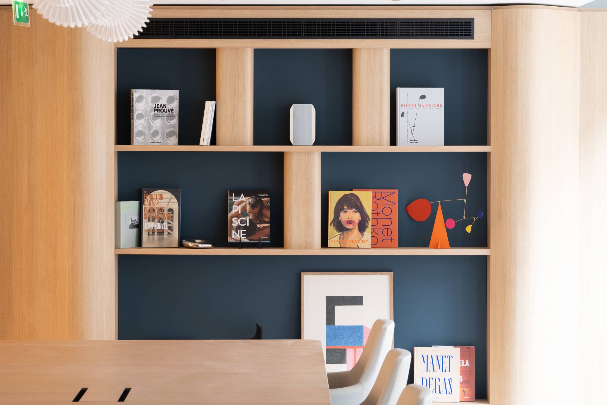 Un plan rapproché sur une bibliothèque bleu nuit intégrée, présentant des ouvrages d'art et un mobile coloré au-dessus d'une table de réunion.