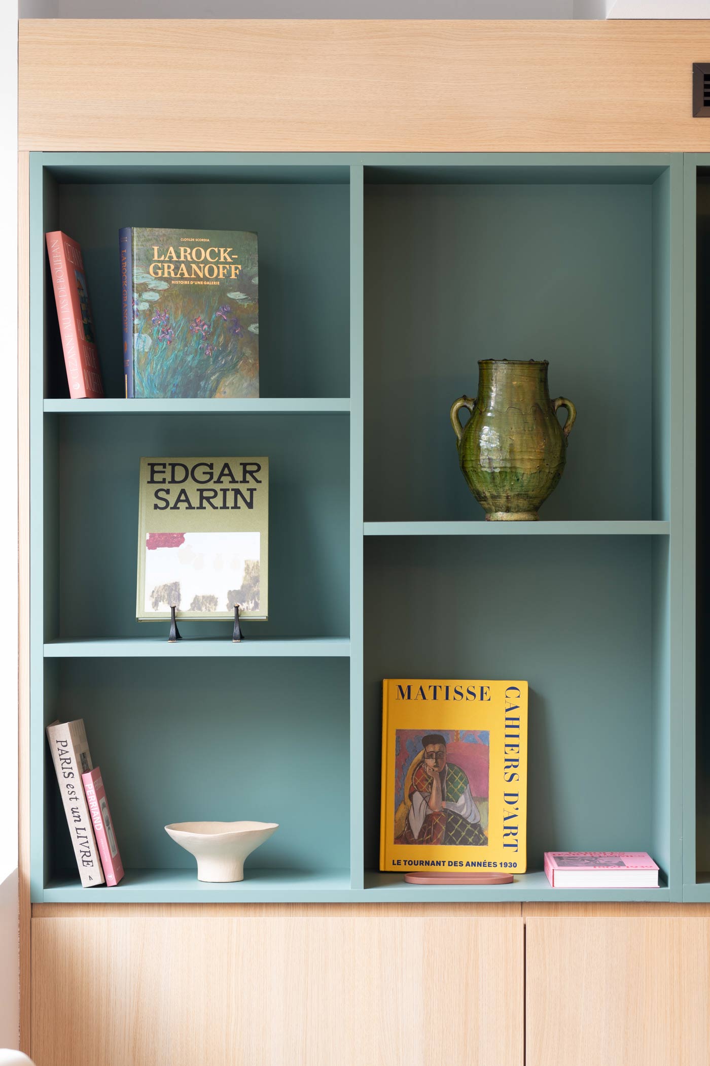 Une section de bibliothèque vert sauge mettant en avant des livres sur Cézanne et Matisse ainsi qu'un vase en céramique vernissée.
