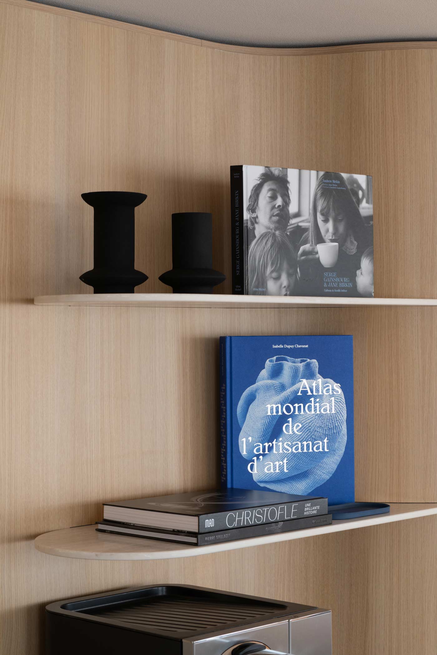Un gros plan sur des étagères en pierre intégrées dans une paroi en bois blond courbé, décorées de vases noirs mats et de livres d'art élégants.