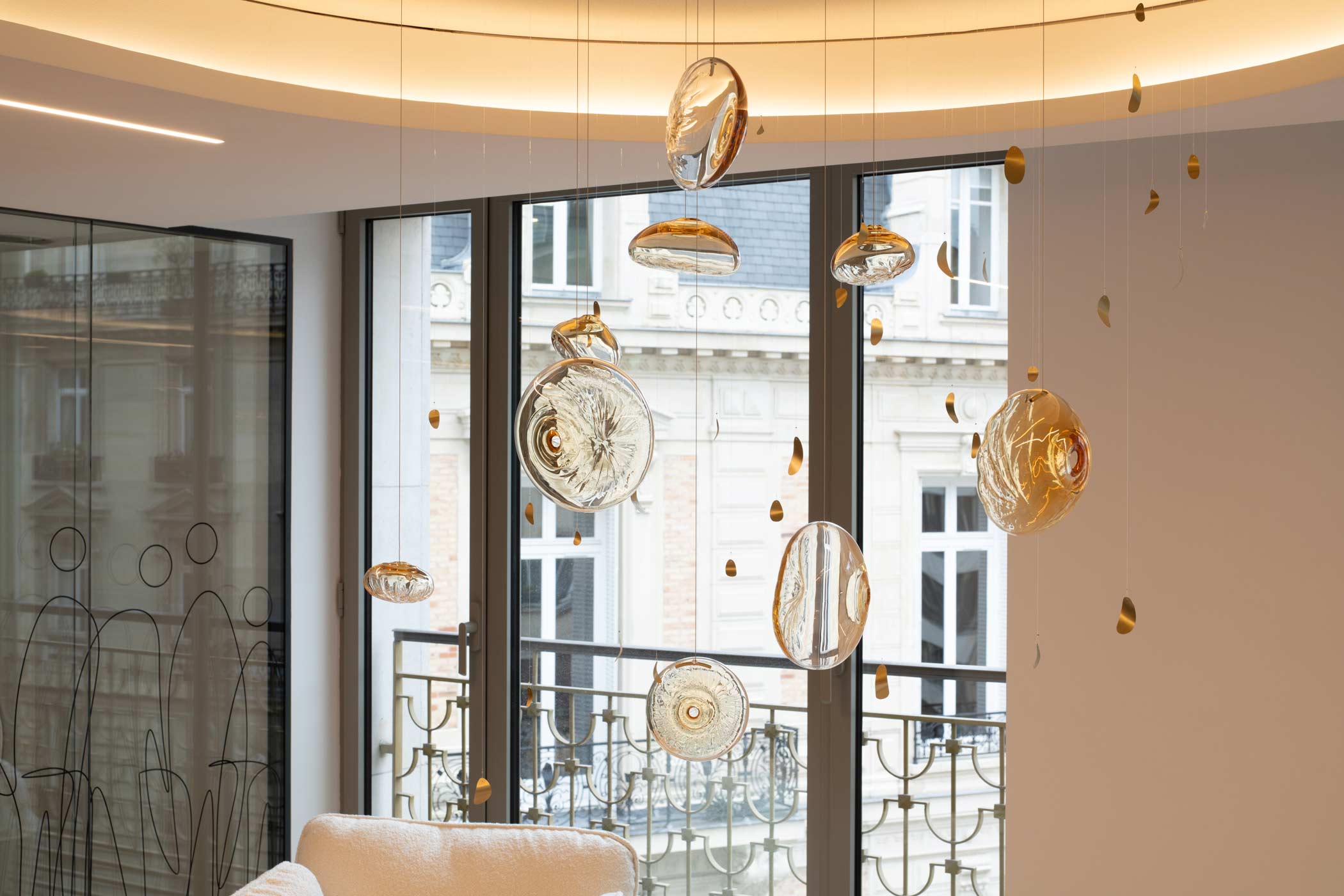 Une vue sur une suspension artistique composée de galets de verre ambré, flottant devant de grandes fenêtres ouvrant sur des façades haussmanniennes.