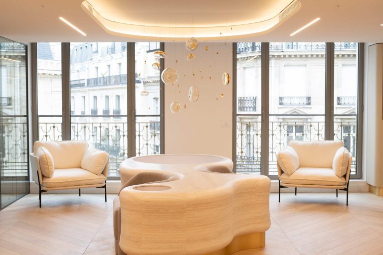 Un salon lumineux mettant en scène une table centrale sculpturale aux formes organiques, entourée de fauteuils blancs et d'un luminaire aérien dans un cadre parisien raffiné.