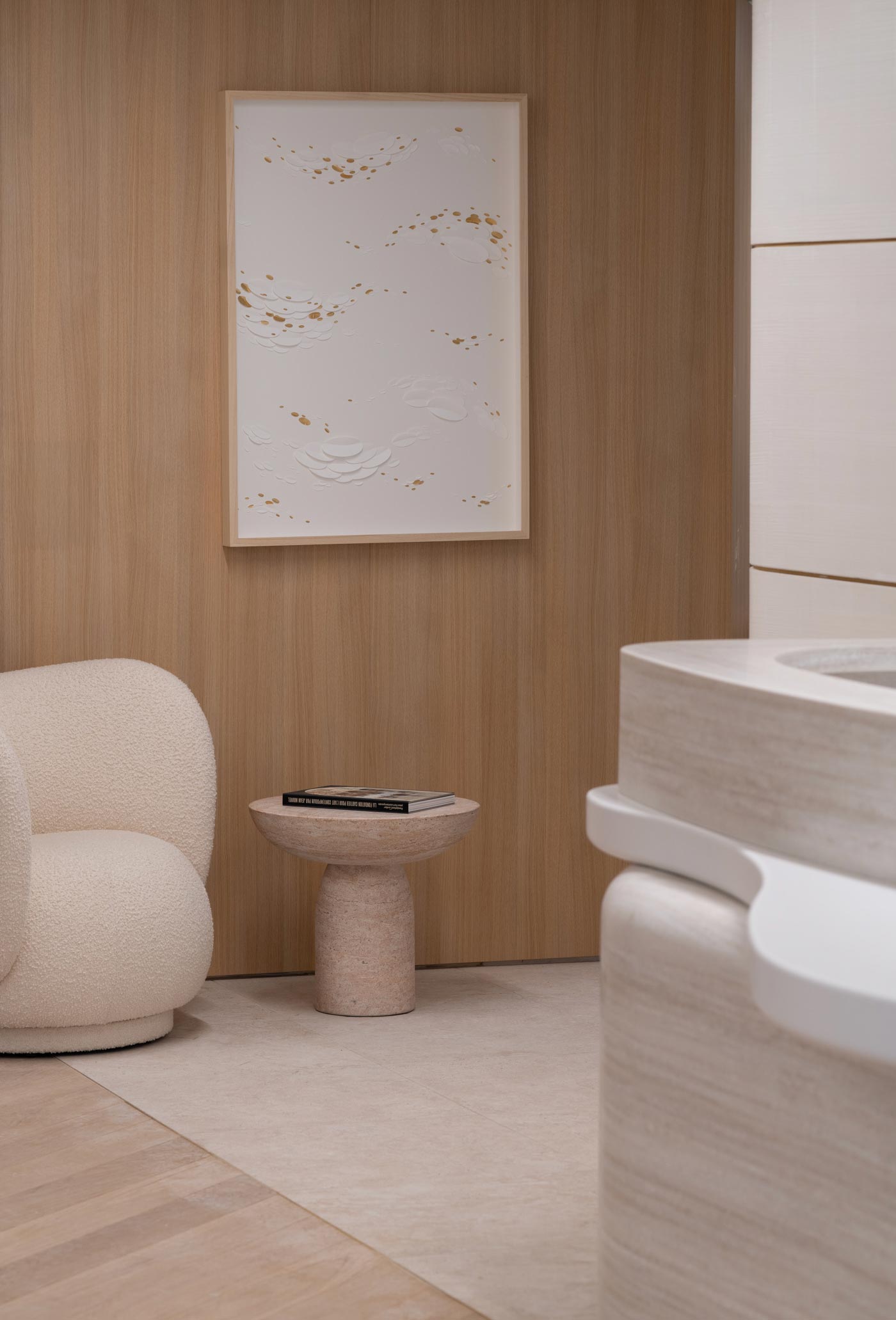 Un coin lecture épuré composé d'un fauteuil bouclé blanc, d'un guéridon en pierre et d'une œuvre murale texturée sur fond de bois.