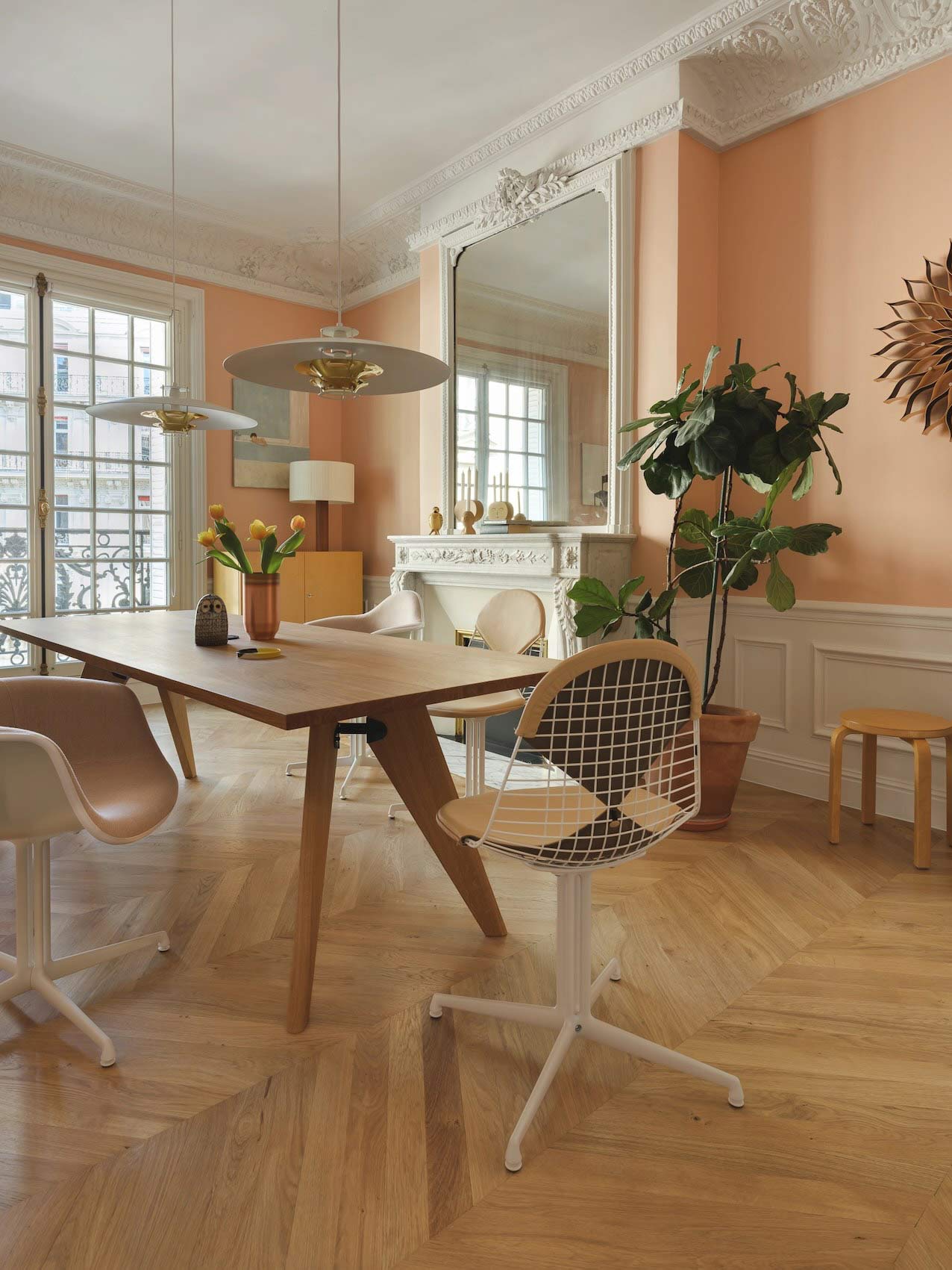 Une salle à manger lumineuse aux murs pêche présente une table en bois entourée de chaises dépareillées sous des suspensions modernes, le tout sur un parquet en chevrons.