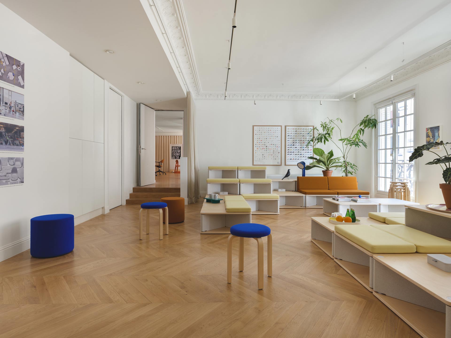 Un espace de travail ouvert et modulable mettant en scène des gradins en bois clair, des tabourets bleus vifs et un parquet en chevrons sous un plafond à moulures classiques.