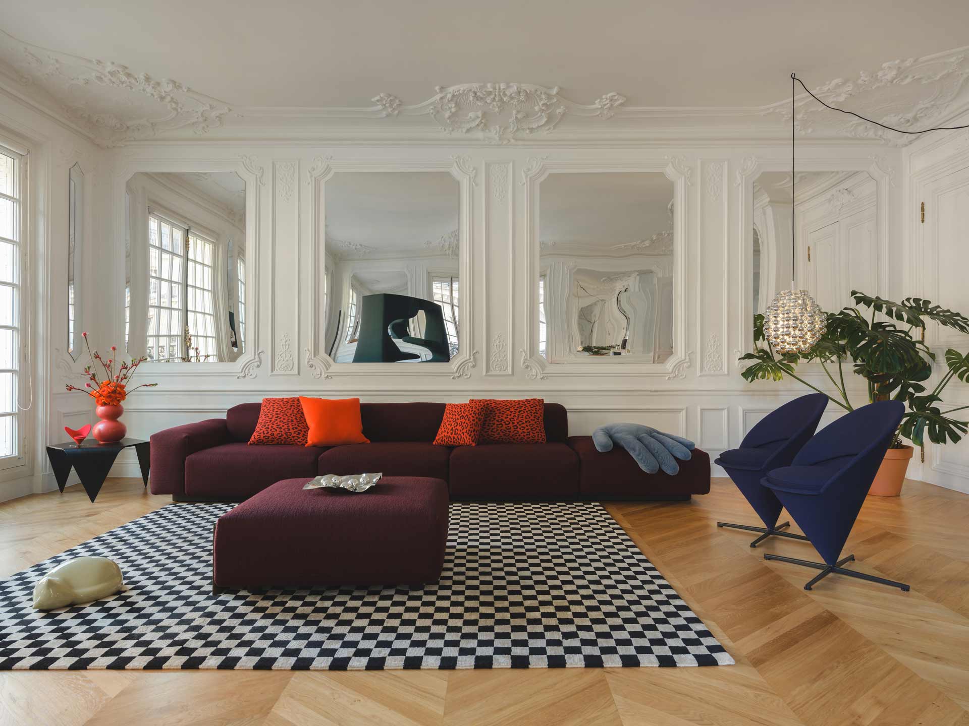 Un salon élégant et audacieux où un canapé lie-de-vin et un tapis en damier noir et blanc font face à un mur orné de boiseries blanches raffinées et de miroirs.