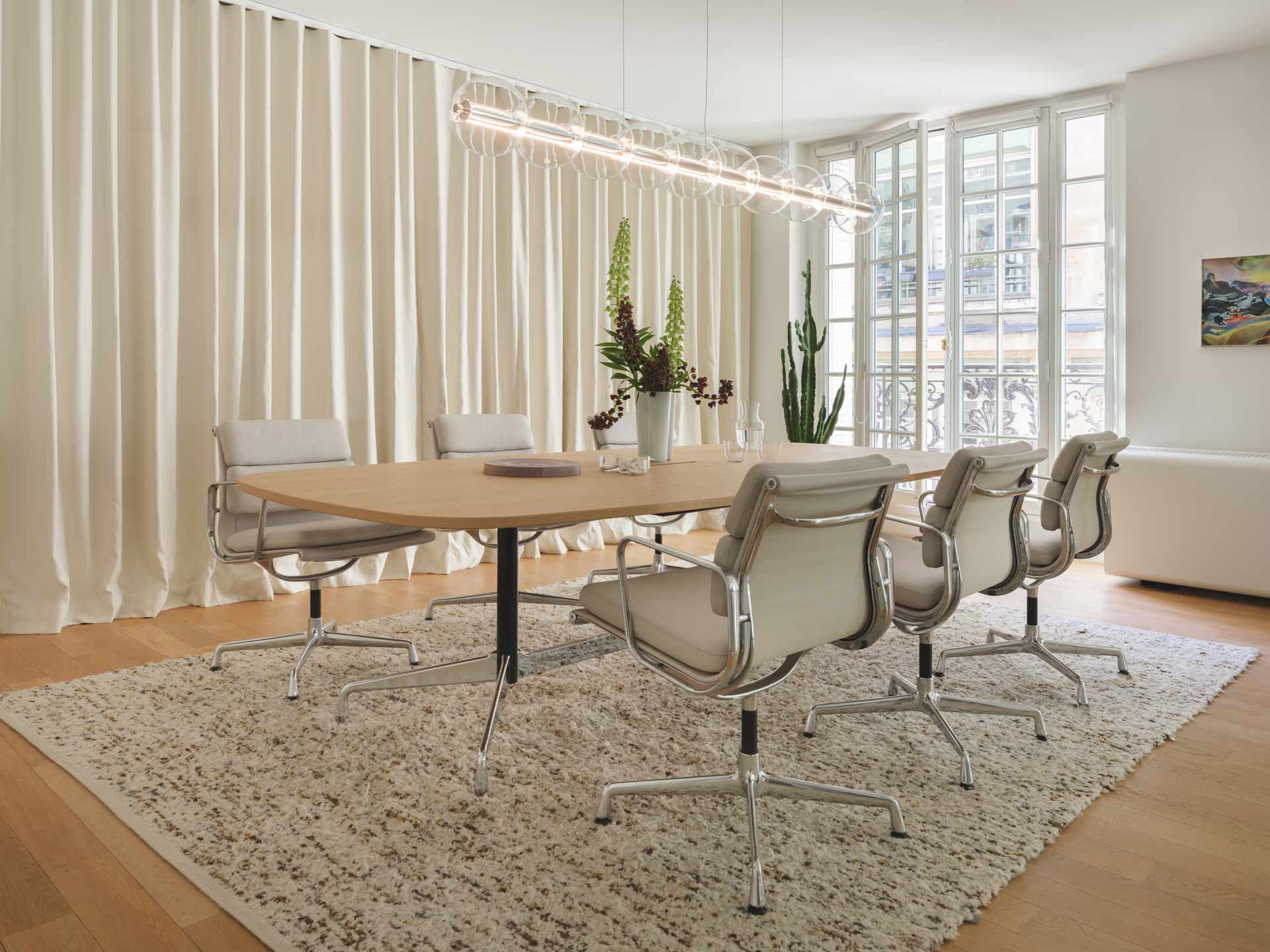 Une salle de réunion lumineuse centrée sur une table ovale en bois entourée de chaises de bureau iconiques, le tout encadré par un rideau drapé et un luminaire tubulaire design.