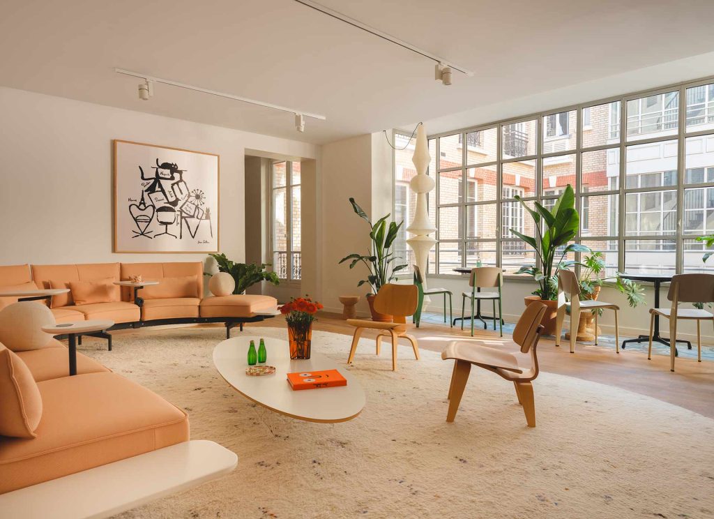 Un vaste espace de détente baigné de lumière naturelle, mêlant un grand canapé incurvé couleur pêche, des fauteuils en bois courbé et de nombreuses plantes vertes.