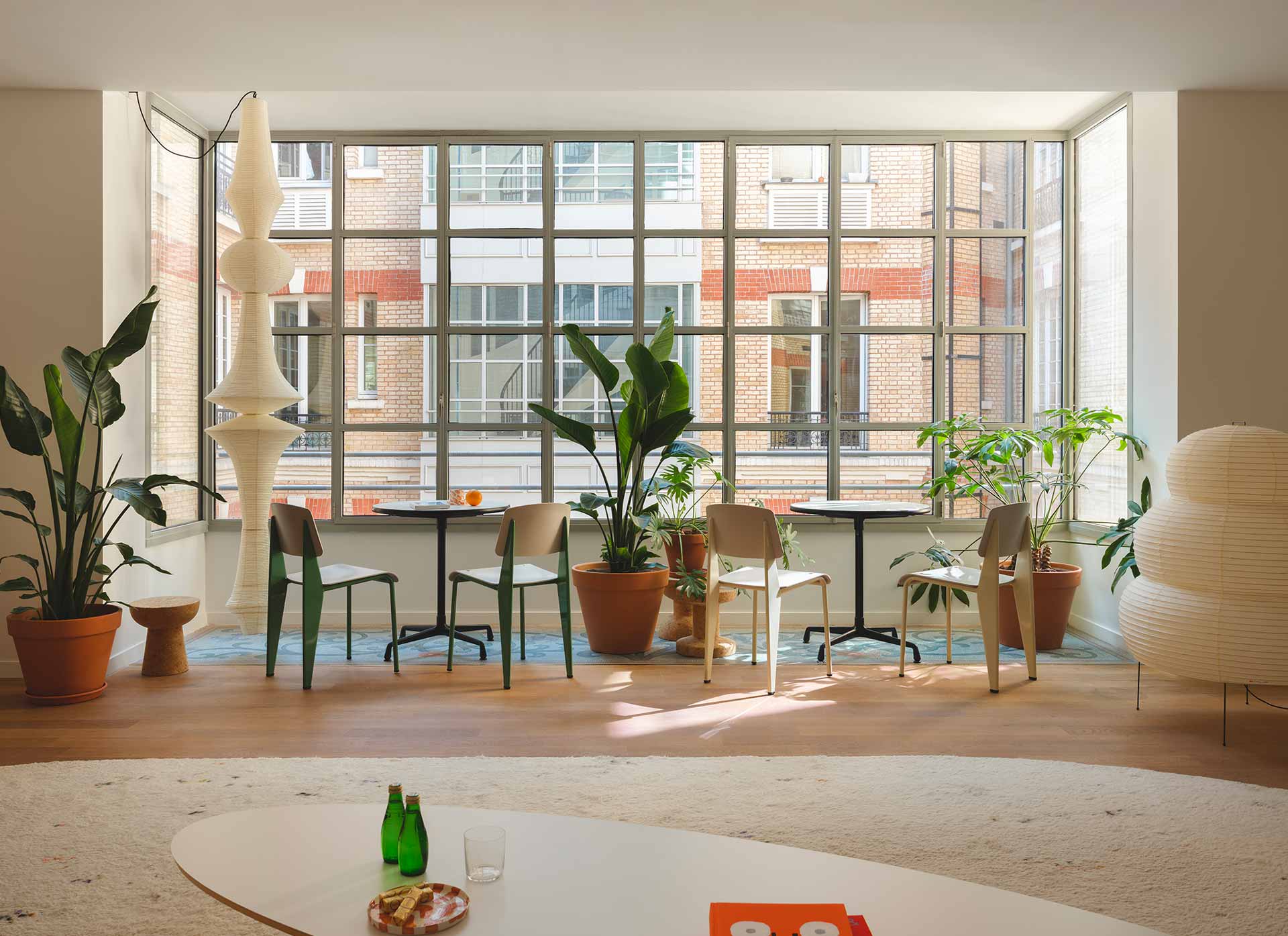 Un coin café convivial aménagé devant une grande baie vitrée quadrillée, agrémenté de tables bistrot, de chaises design et de sculptures lumineuses en papier d'inspiration japonaise.