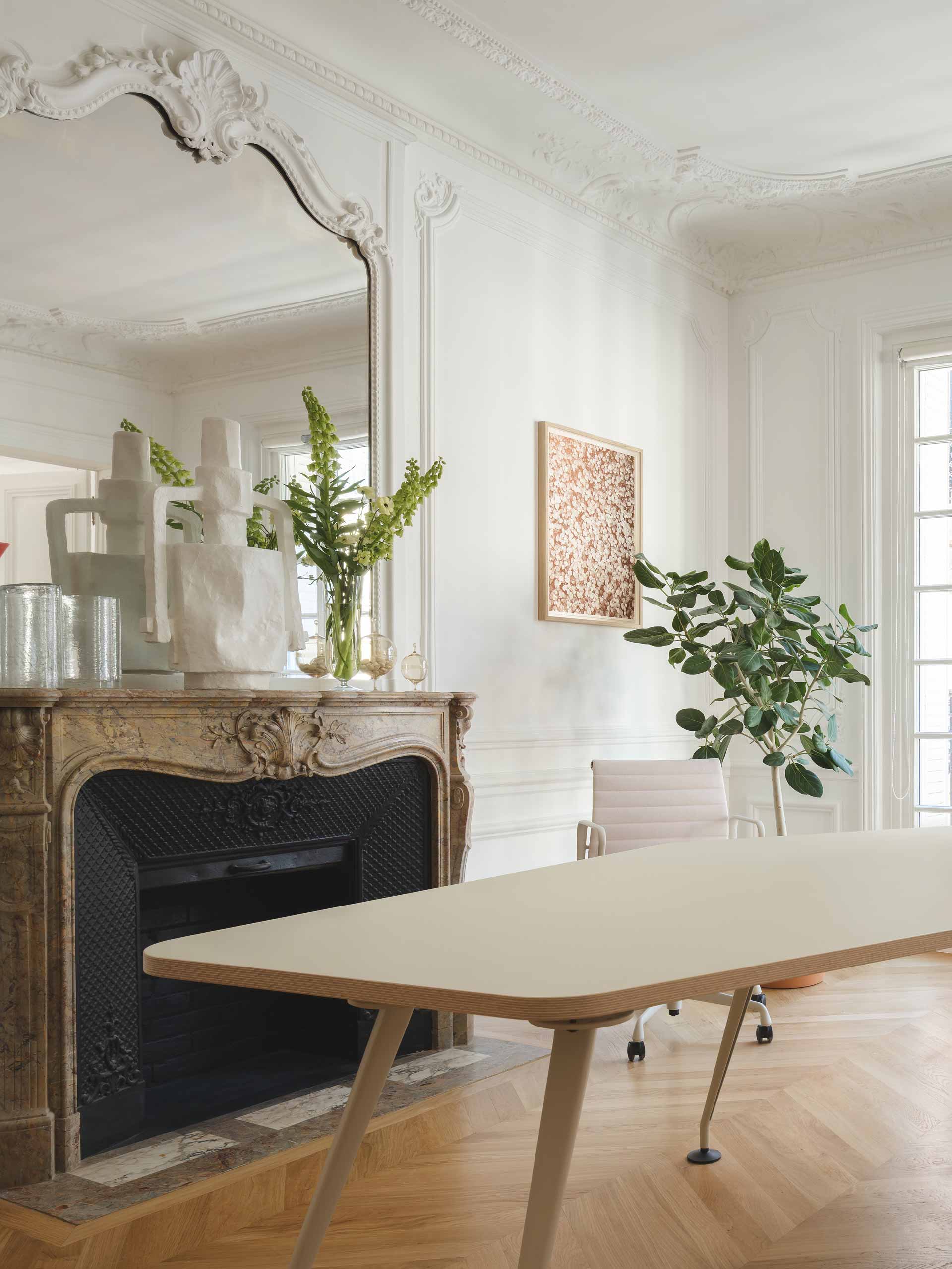 Un bureau élégant et minimaliste est installé dans une pièce classique avec une cheminée sculptée, un grand miroir orné et des moulures blanches raffinées.