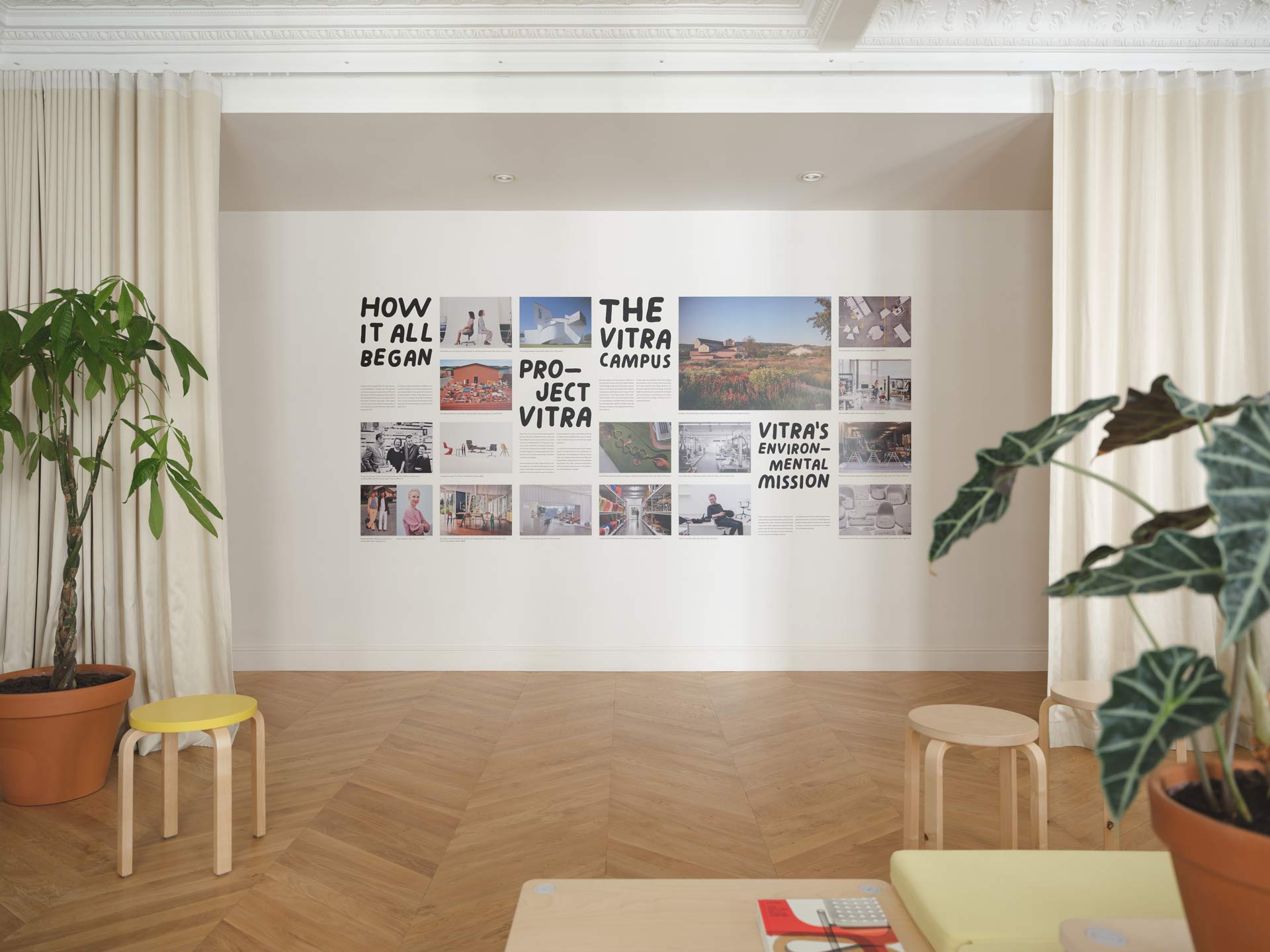 Un mur d'exposition présente l'histoire et les missions de Vitra à travers une grille de photographies et de textes, encadré par des rideaux beiges et des plantes en pot.