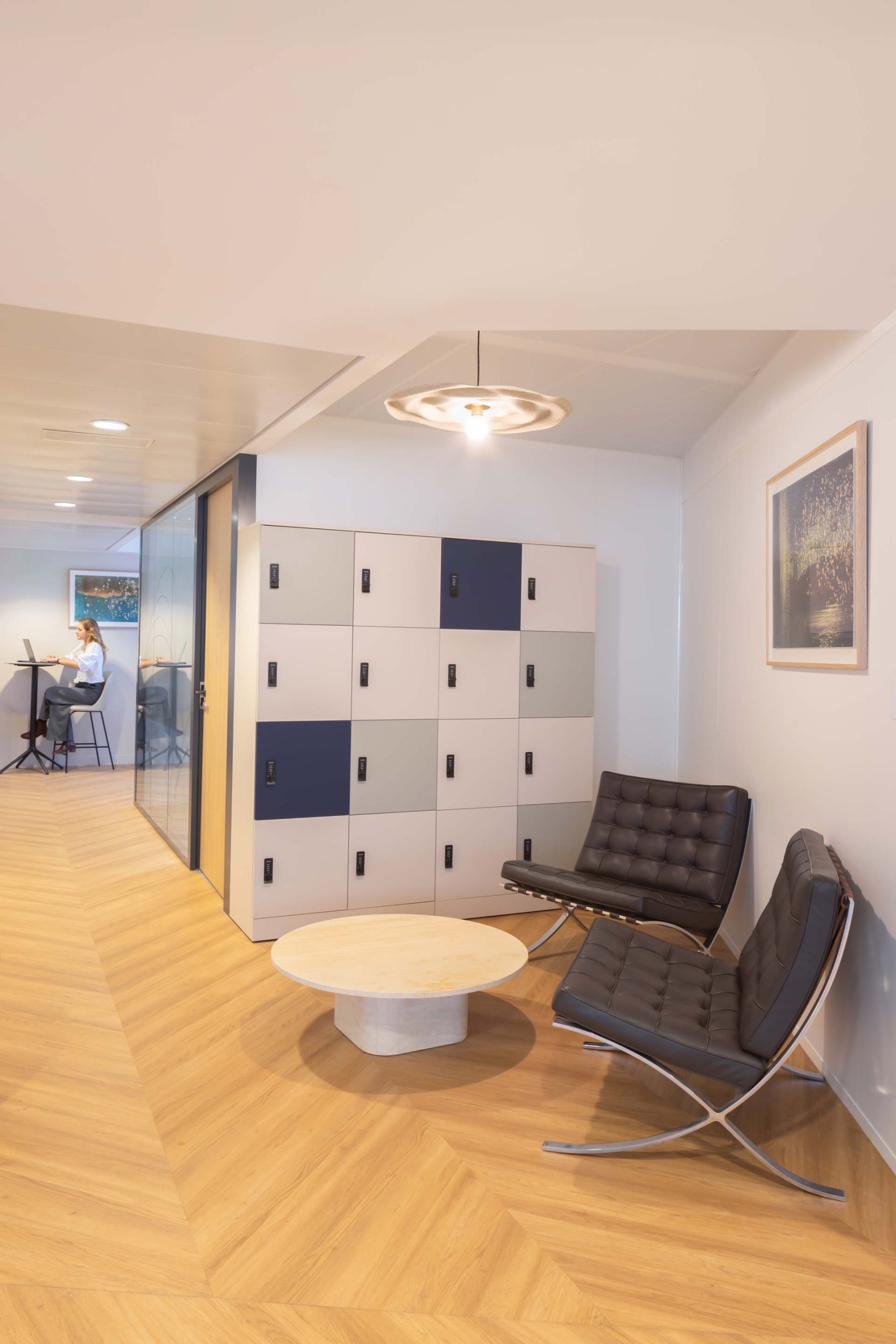 Un couloir de bureau avec des casiers de rangement multicolores (bleu, beige, gris), des fauteuils en cuir noir (style Barcelona) et un sol en bois chaleureux.