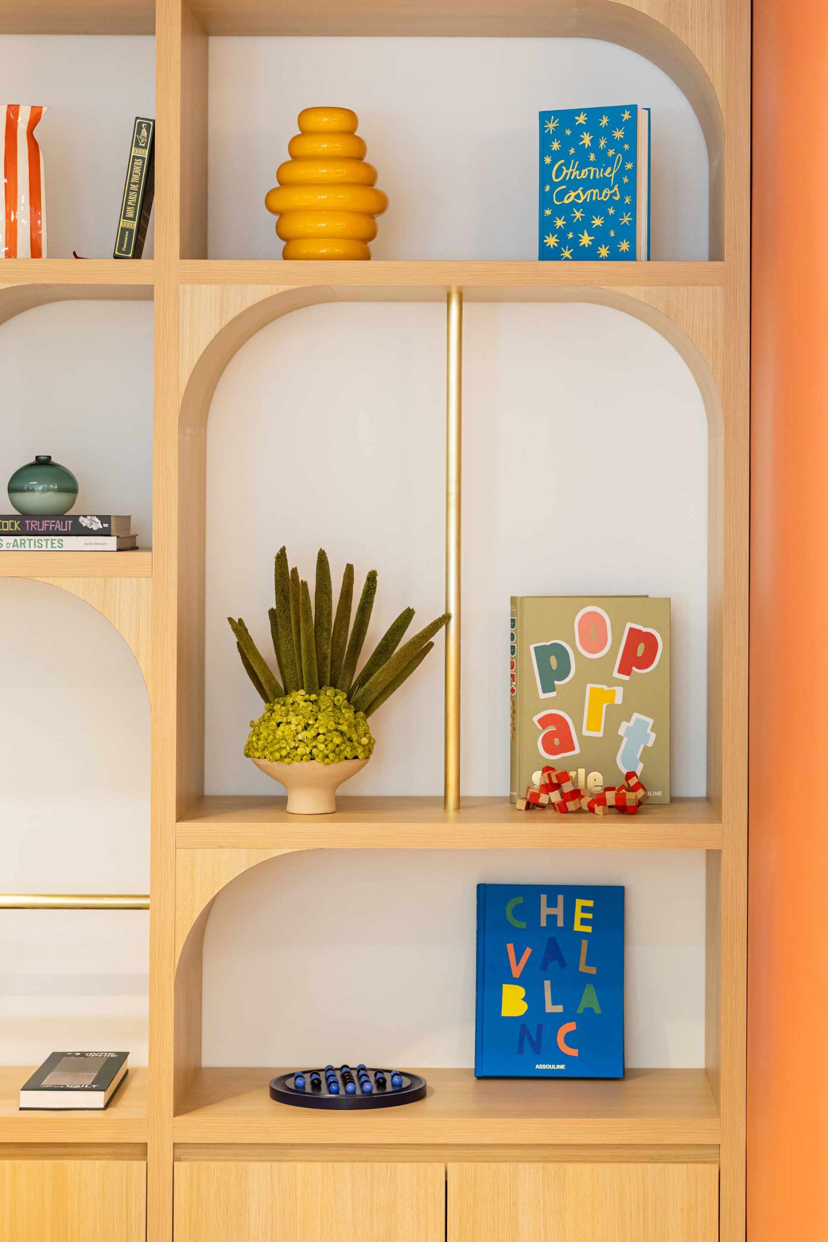 Gros plan sur une étagère en bois aux niches arrondies, décorée avec soin : on y voit des livres d'art colorés (Pop Art, Cheval Blanc), un vase jaune en forme de ruche et une petite plante grasse.