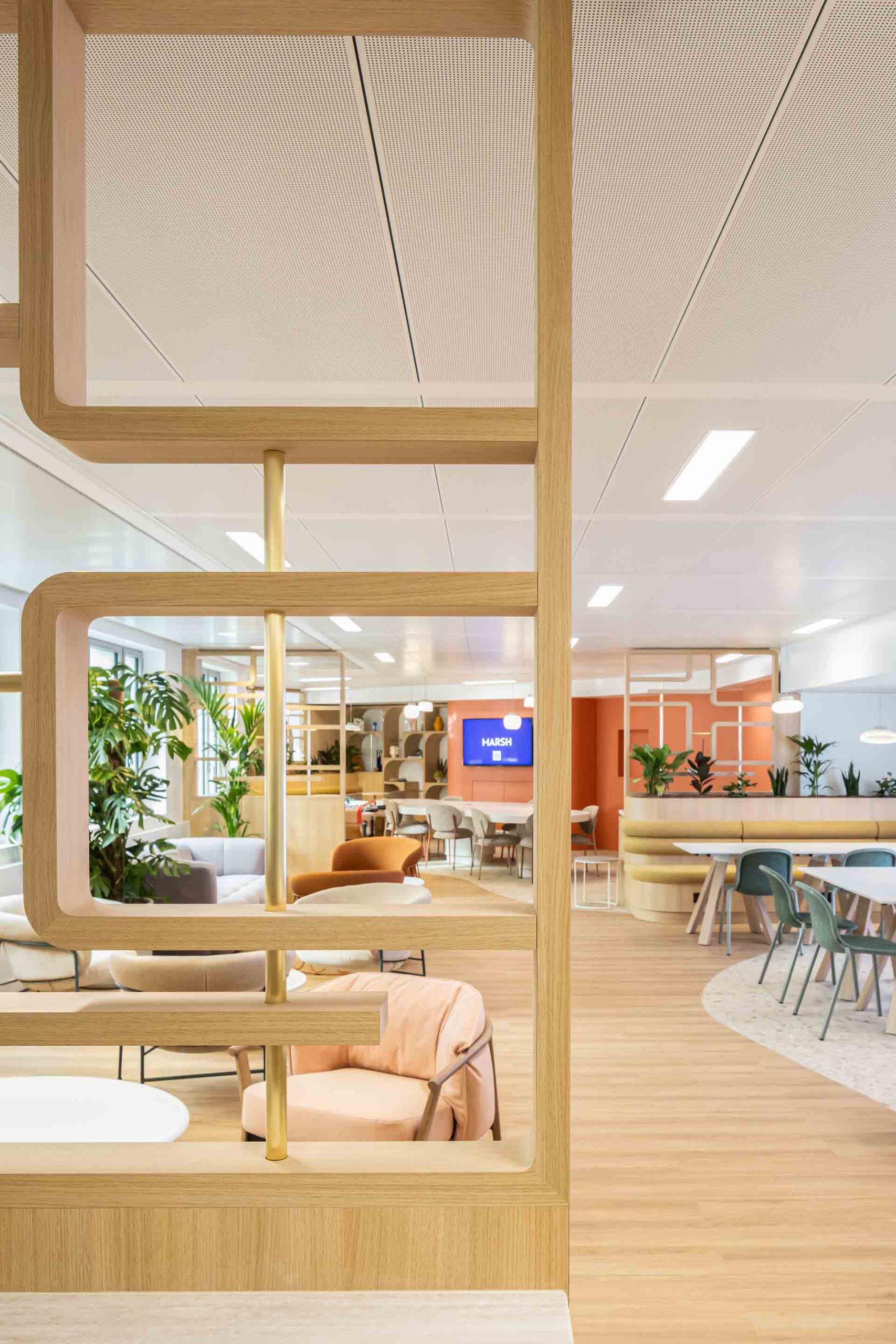 Vue à travers une cloison décorative en bois ajouré montrant un salon lounge avec des fauteuils confortables aux tons pastel (rose, orange, gris) et de nombreuses plantes vertes.