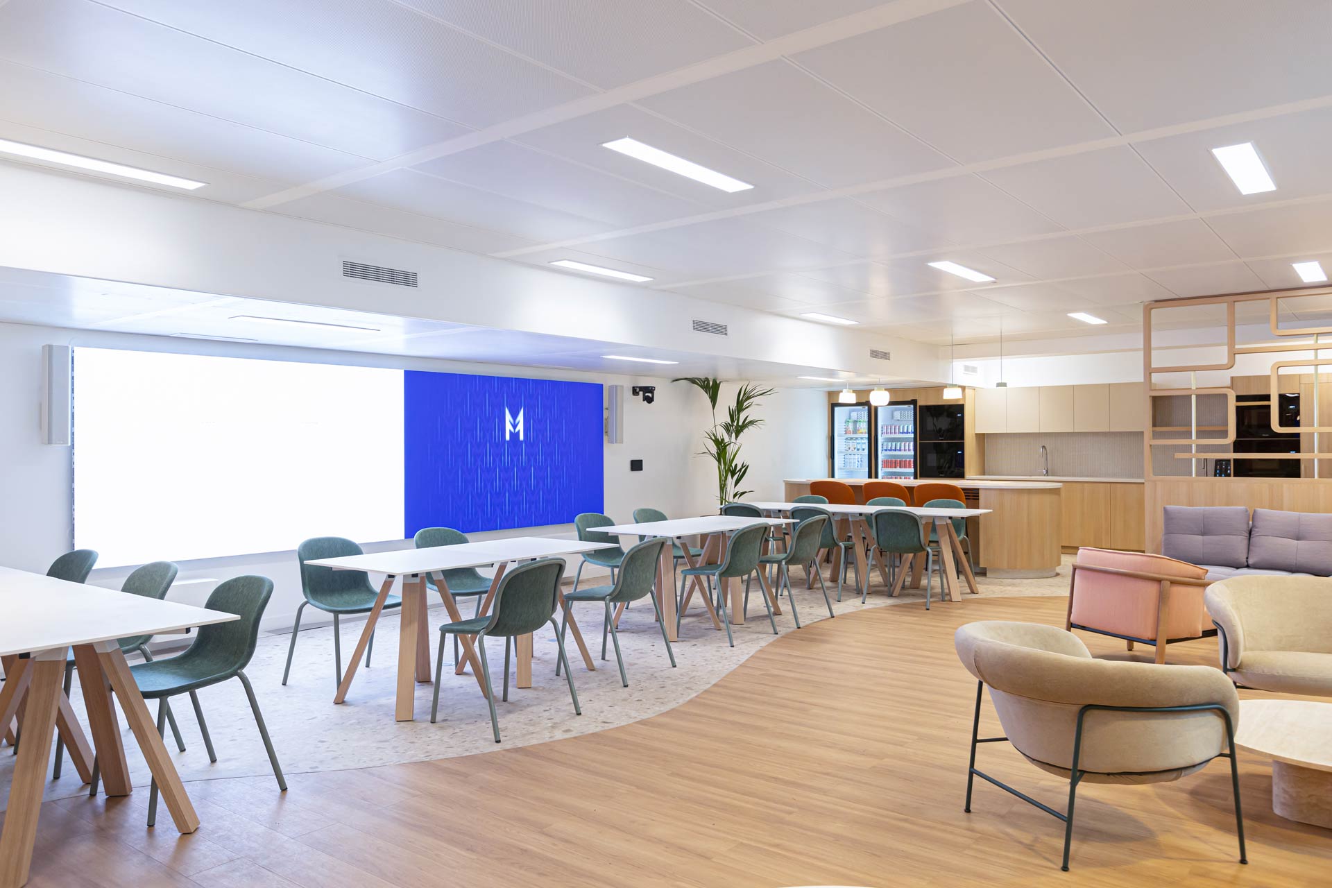 Espace polyvalent de restauration ou de travail avec plusieurs tables blanches et chaises vertes, ainsi qu'un grand mur d'écrans lumineux bleus et une cuisine équipée en arrière-plan.