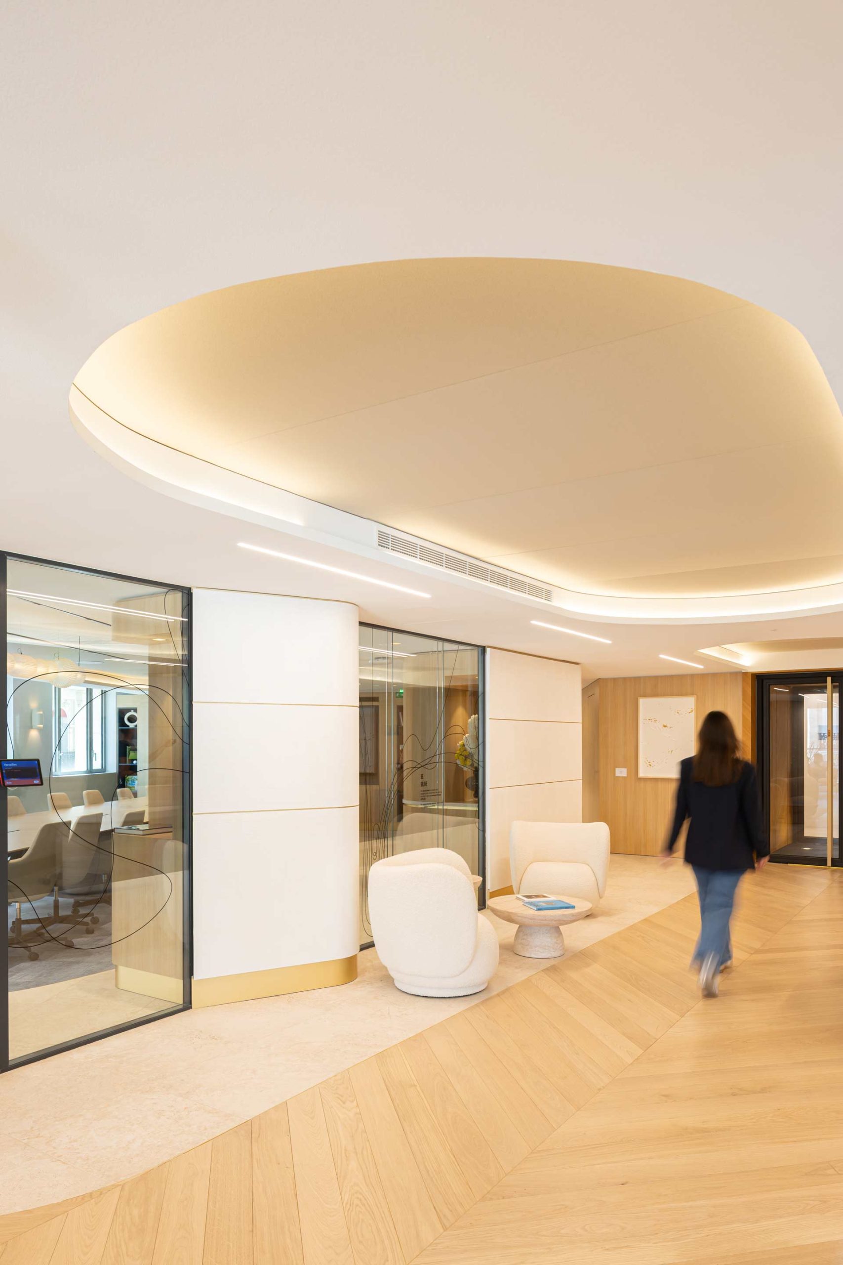 Vue d'un couloir élégant avec un plafond à caisson lumineux aux formes fluides, des fauteuils blancs arrondis et une paroi vitrée laissant entrevoir une salle de réunion.