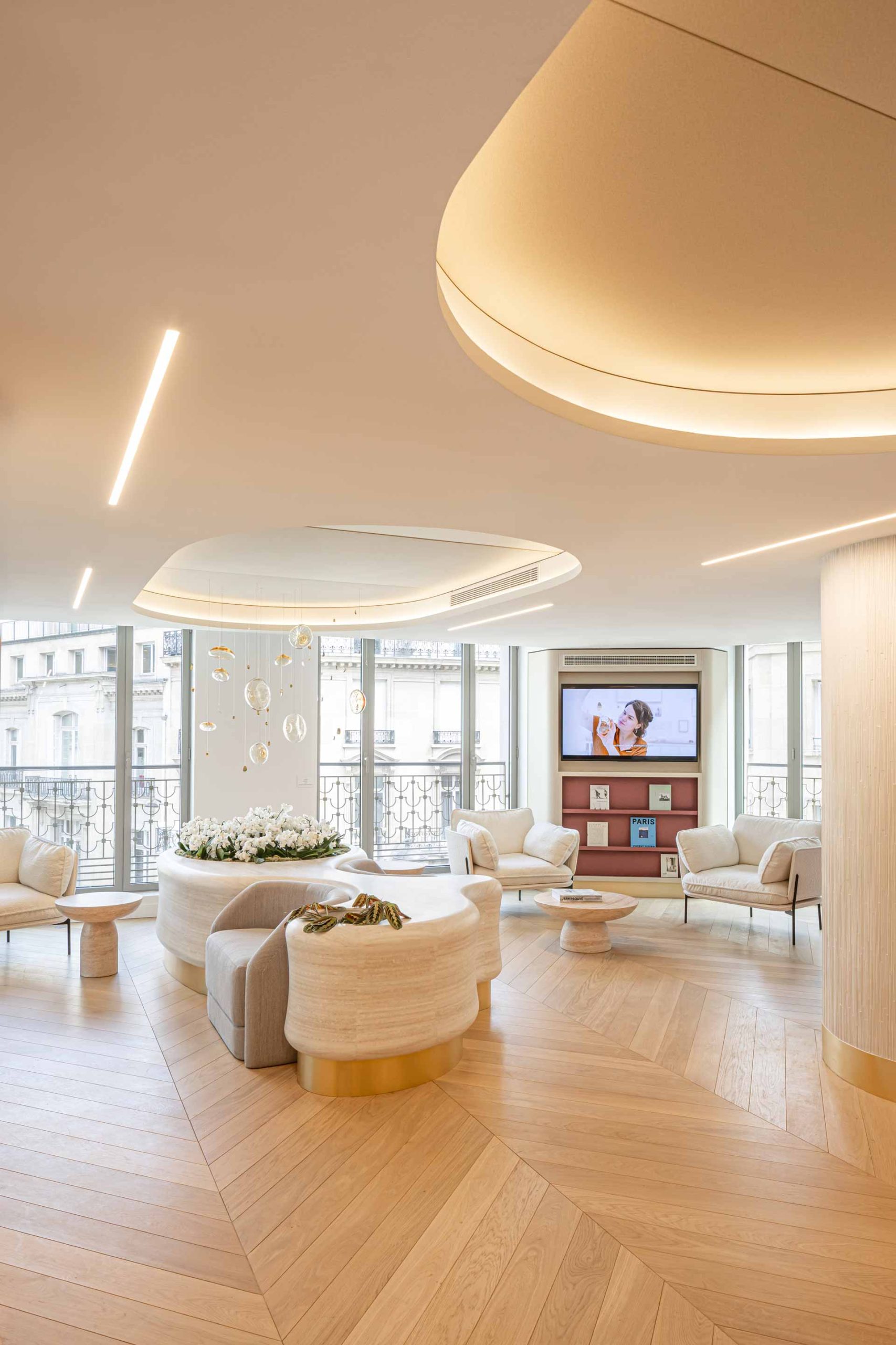 Un salon d'attente lumineux avec des fauteuils blancs design, une table basse en pierre et un écran plat intégré dans un meuble de rangement aux tons bordeaux.