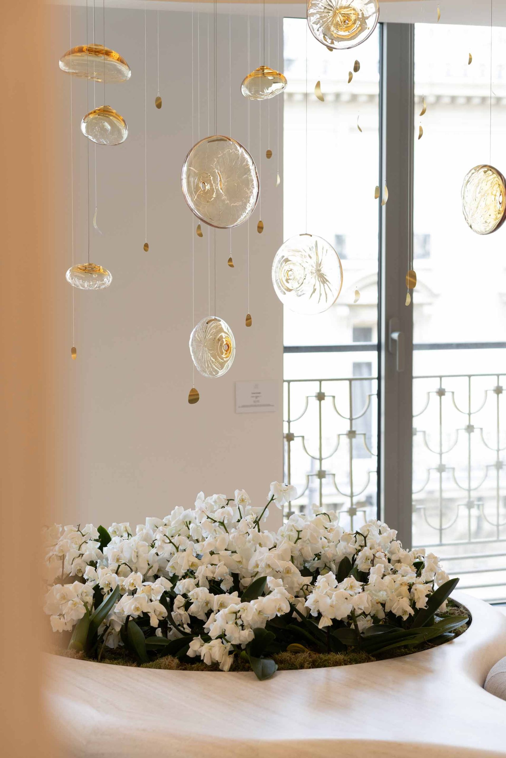 Gros plan sur une décoration florale composée d'orchidées blanches surmontée d'une installation artistique de suspensions en verre soufflé devant une fenêtre.