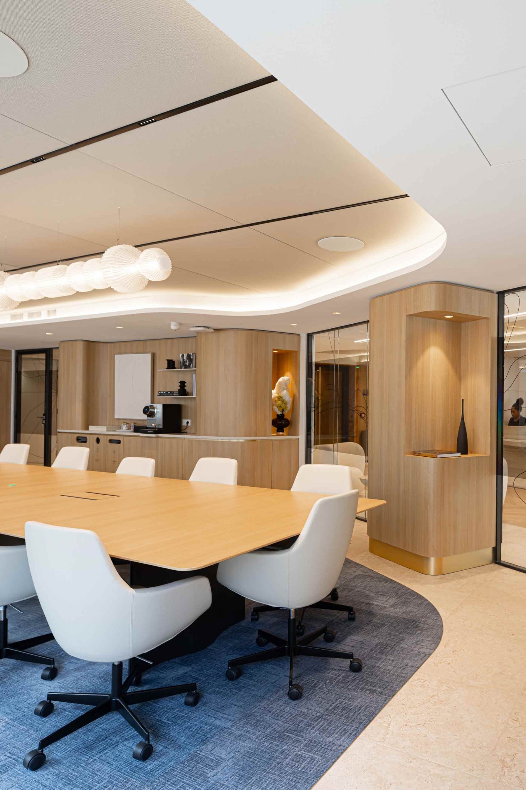 Vue partielle d'une salle de conférence avec une grande table en bois clair, des fauteuils de bureau blancs sur un tapis bleu et des finitions murales en bois blond.