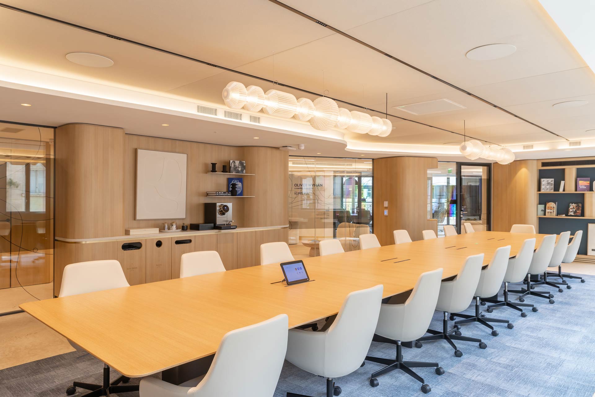 Vue d'ensemble de la longue salle de conférence montrant l'alignement des chaises blanches, un luminaire design suspendu de forme organique et un coin café intégré.
