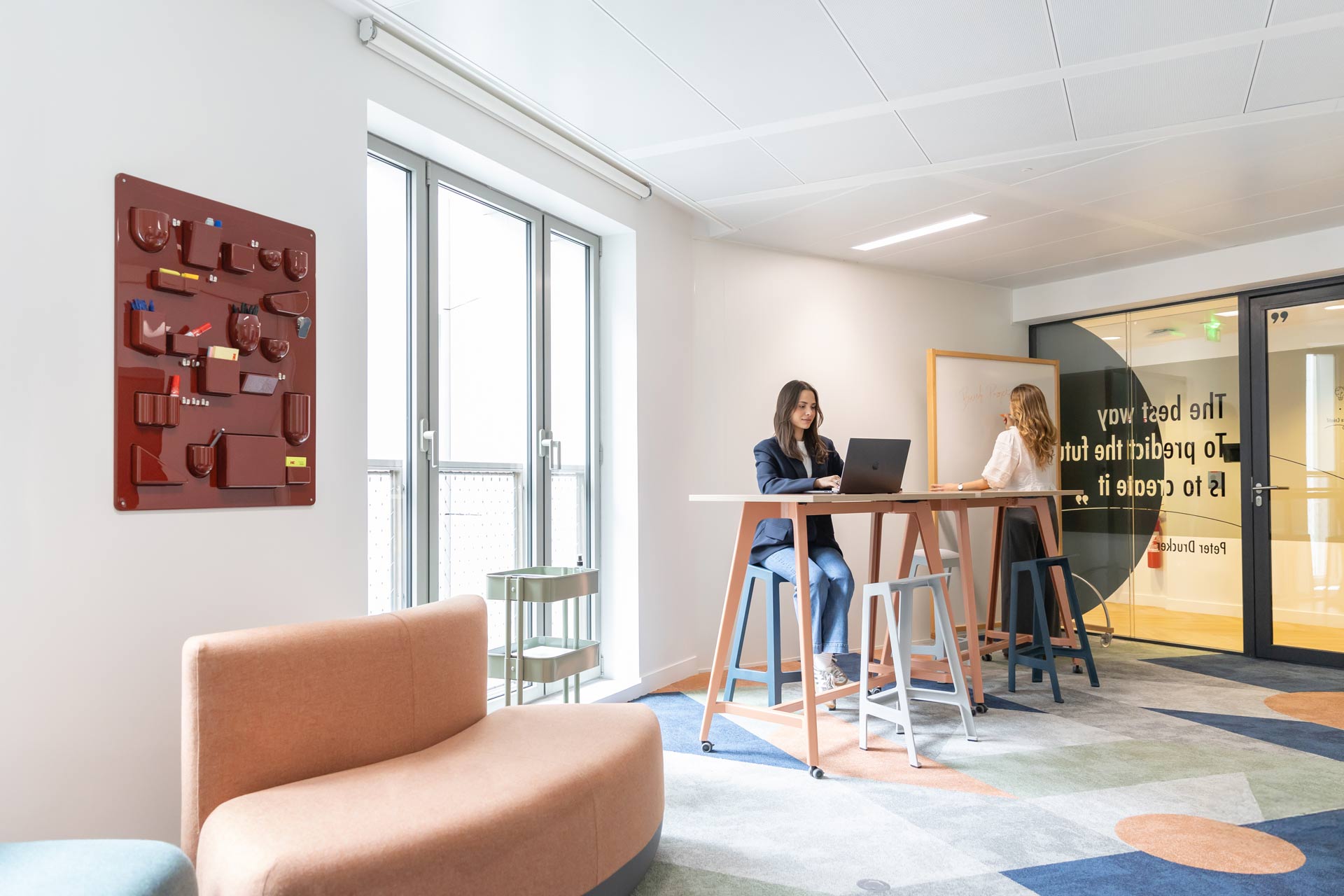 Un espace de travail collaboratif appelé "Creative Room" comprenant une table haute, des tabourets colorés, un tapis géométrique et un panneau de rangement mural rouge.