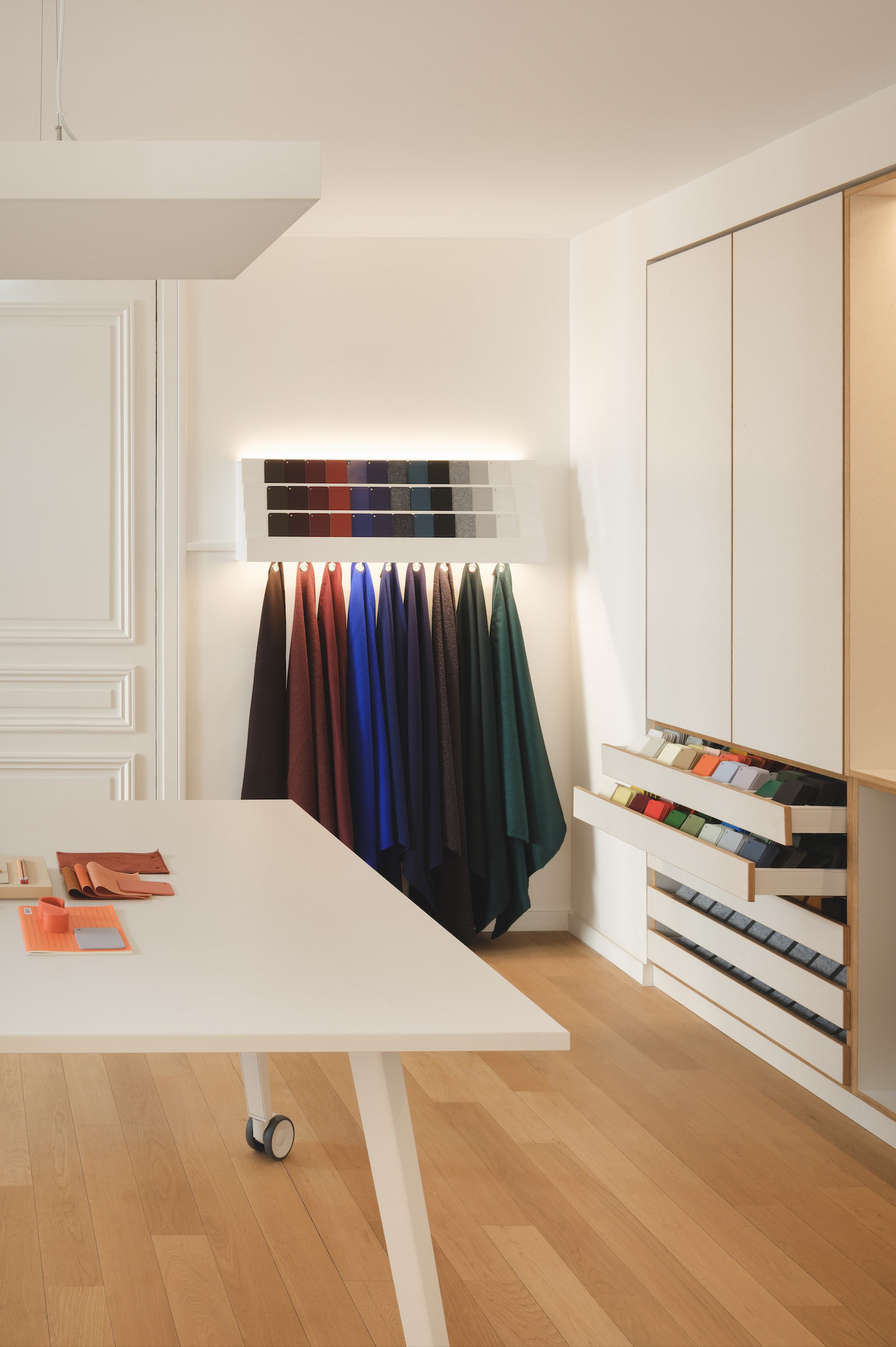 Un atelier de design lumineux expose une vaste matériauthèque avec des échantillons de tissus colorés suspendus et des nuanciers disposés sur une grande table blanche.