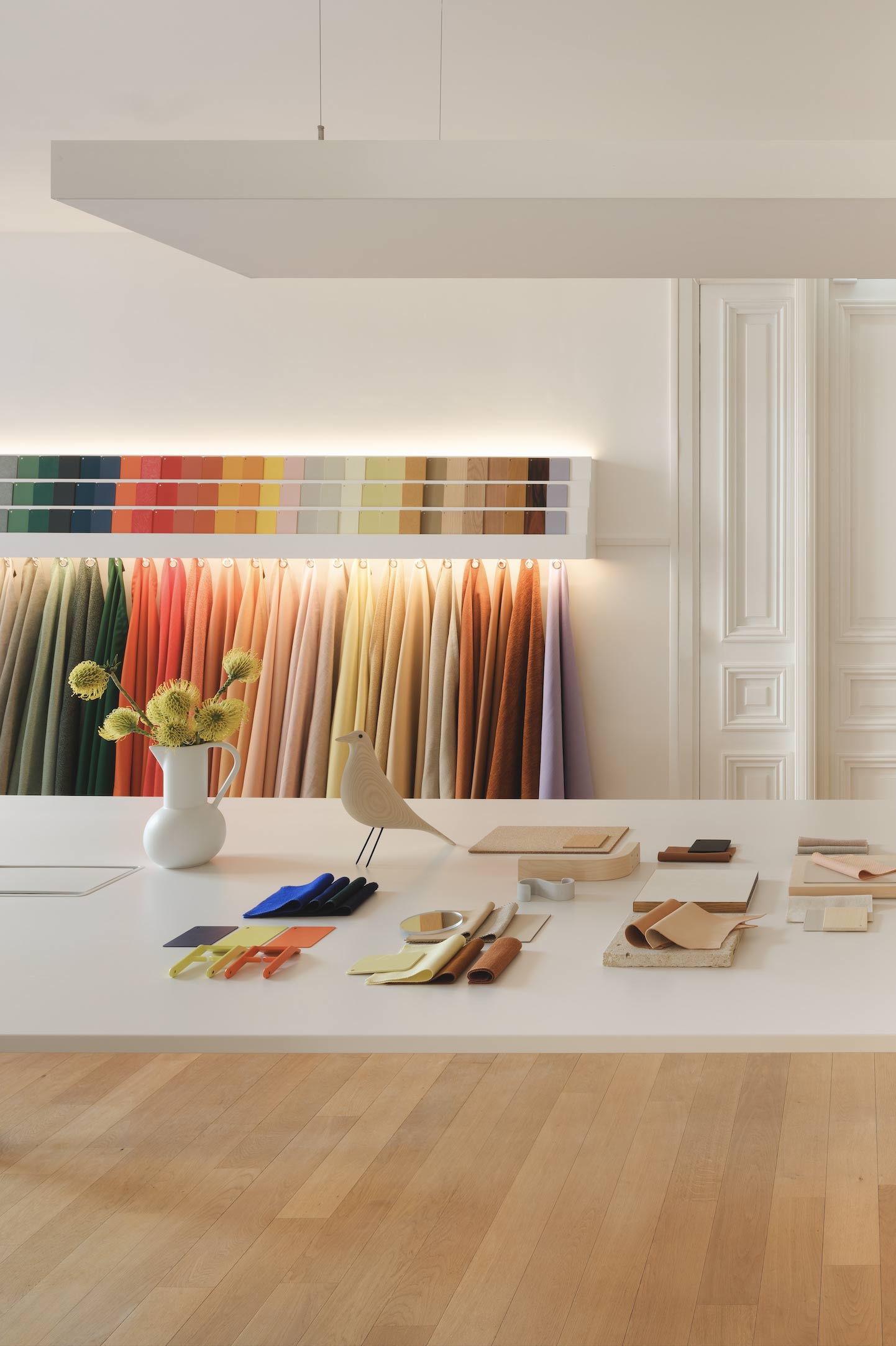 Un atelier de design lumineux expose une vaste matériauthèque avec des échantillons de tissus colorés suspendus et des nuanciers disposés sur une grande table blanche.